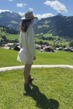 Woman in a white dress and hat standing on a grassy field with mountains and a village in the background.